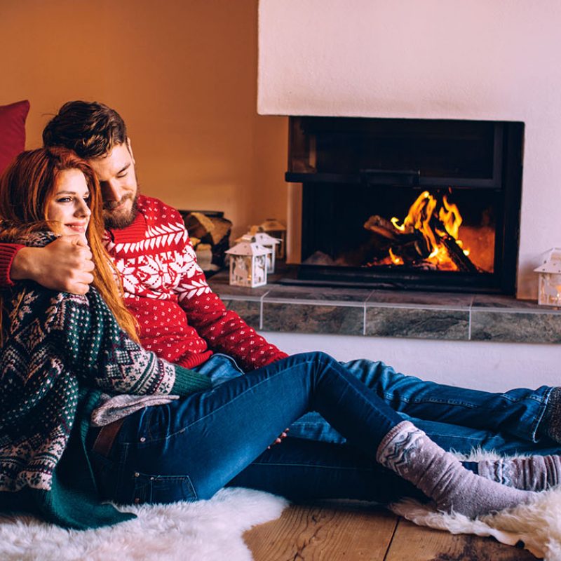 Couple on vacation at mountain cabin. Sitting on the floor on a fur by fireplace in cozy  living room on Christmas. Wearing festive knitted sweaters and socks. Austrian Alps.